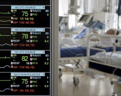 In the foreground, a monitor showing heart rates and other vital signs of four people. In the background, patients in hospital beds.