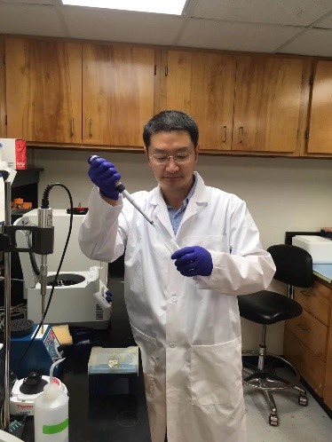 A scientist wearing a lab coat and holding a pipette in front of a workbench with scientific instruments.