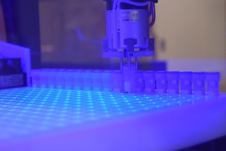 Many small, plastic vials, one of which a robot arm is lifting from an illuminated tray.