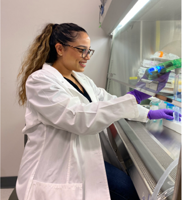Jade, wearing a white lab coat and purple latex gloves, holds a pipette aid and tube inside a biosafety cabinet.
