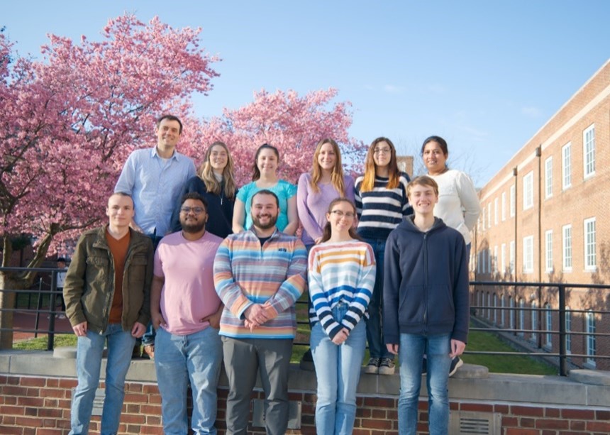 Eleven people standing outside side-by-side in two rows near a campus building.