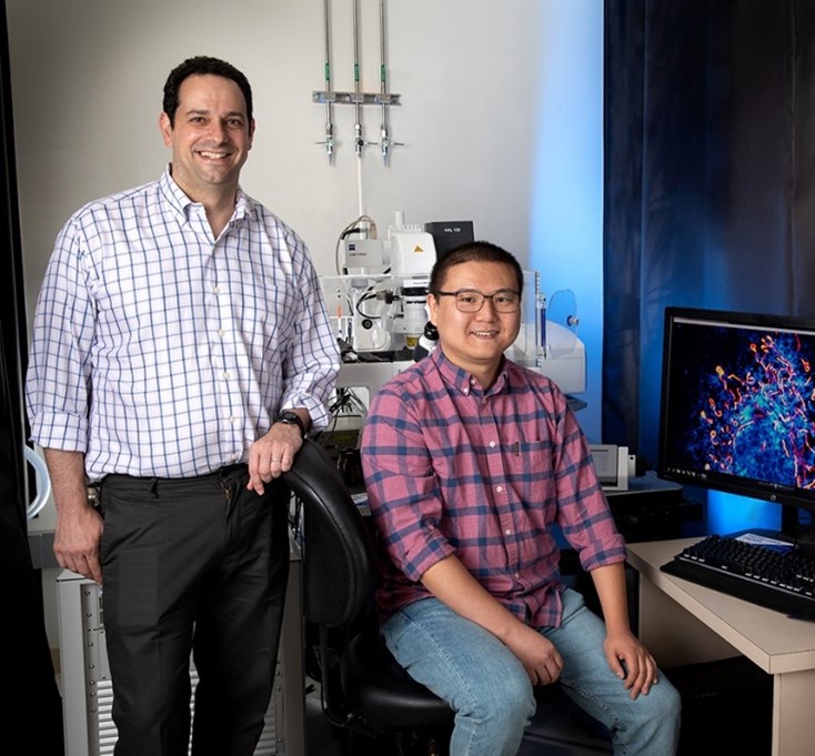 Dr. Saghatelian standing beside a seated Dr. Chu in the laboratory. Beside them is a computer, and behind them is a microscope.