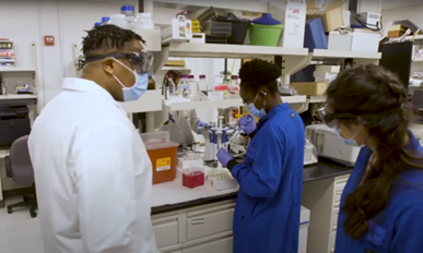 Bryan, a student, and Dr. Akinola standing in the lab, wearing lab coats, gloves, surgical face masks, and safety glasses. Dr. Akinola uses a pipette at the lab bench.