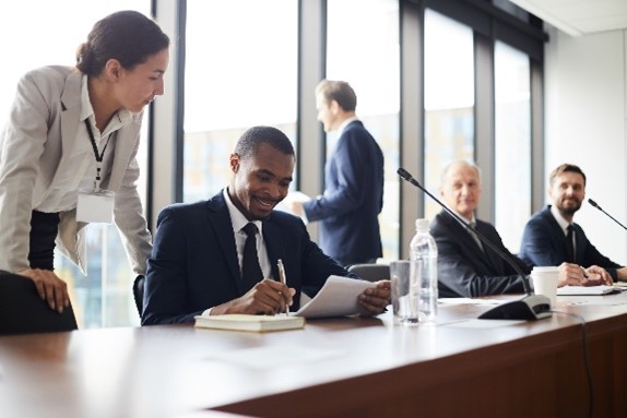 Four people at a long conference table while a fifth person stands by a window. Two are looking at a document together.